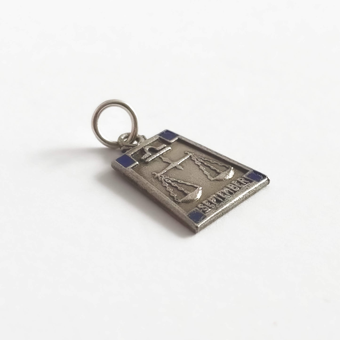 Silver trapezoid shape pendant with Libra zodiac sign and 'September' text with four dark blue enamel corner squares, on a white background