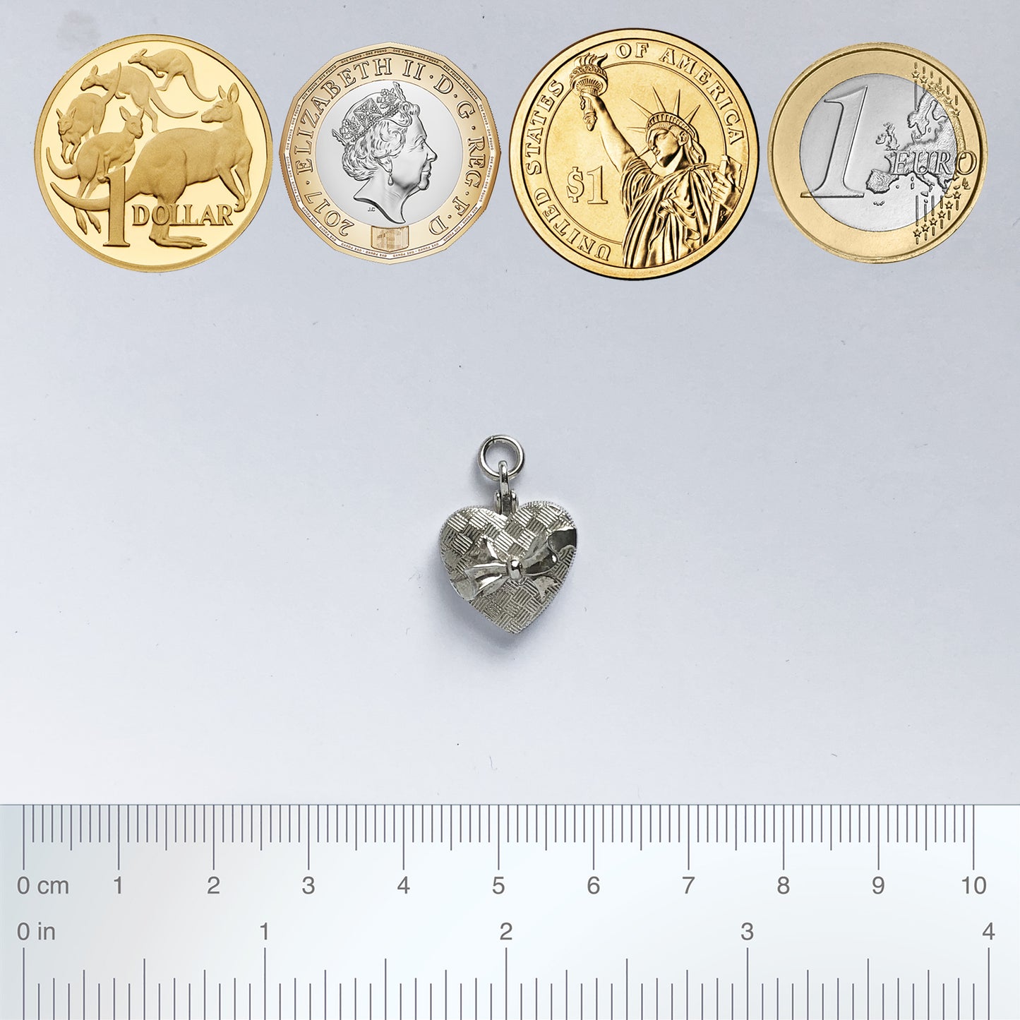silver heart shaped chocolate box charm with coins and ruler for scale on a grey background