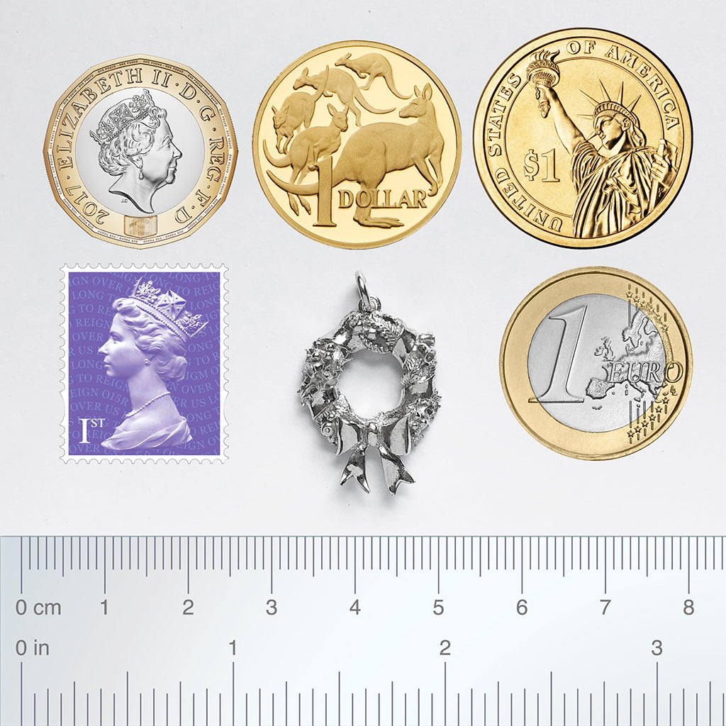 silver Christmas wreath charm with coins stamp and ruler for scale on a light grey background