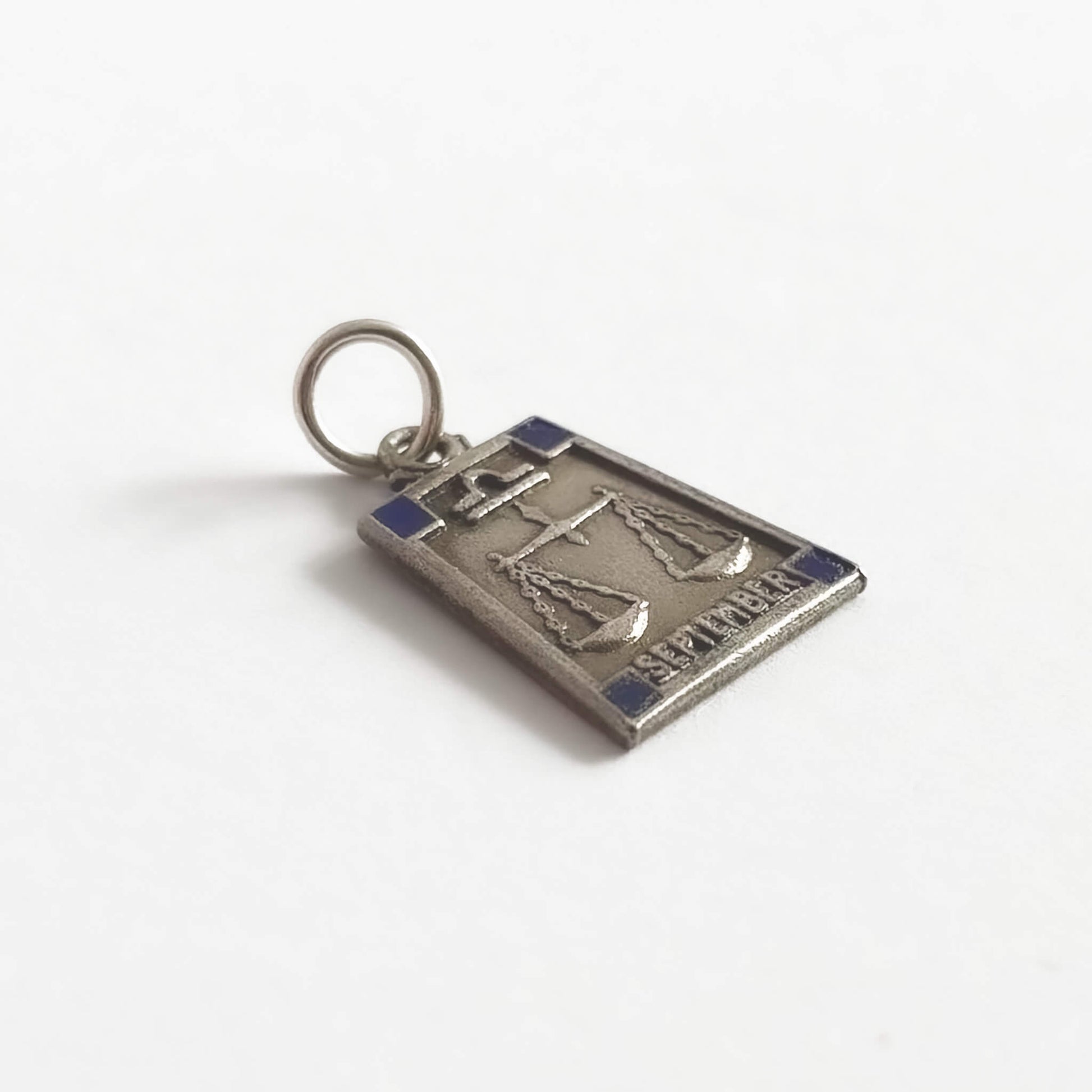 Silver trapezoid shape pendant with Libra zodiac sign and 'September' text with four dark blue enamel corner squares, on a white background