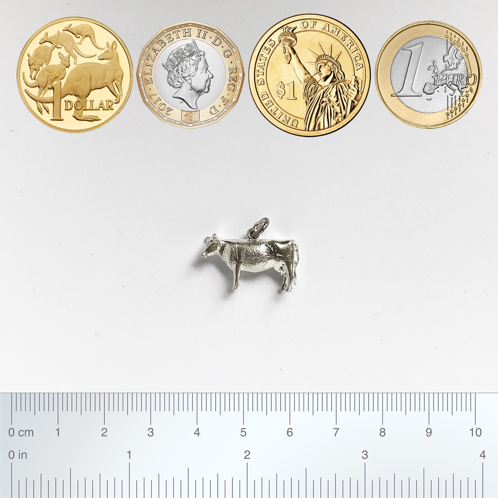 Sterling silver cow charm with coins and ruler for scale on a grey background