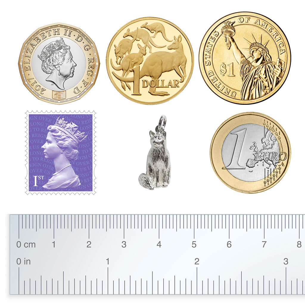 A sitting silver Birman cat charm with coins, a postage stamp and a ruler for scale, all on a white background.