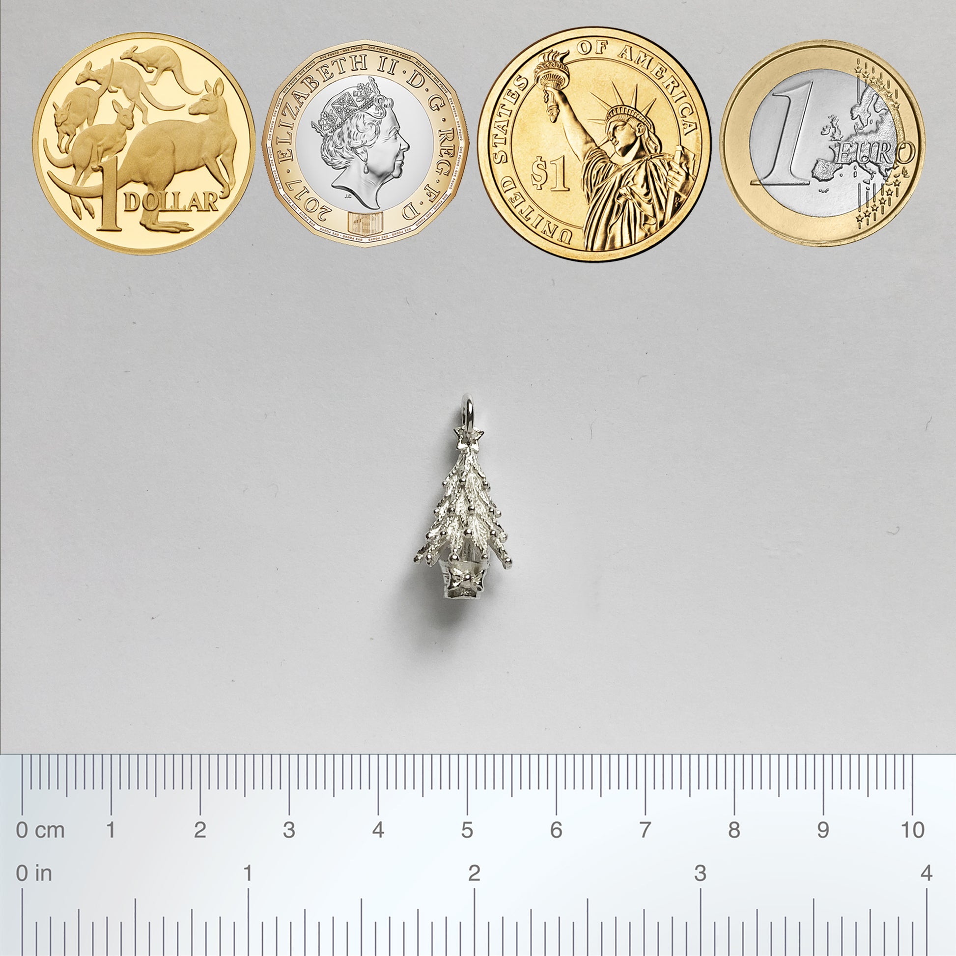 silver Christmas tree charm with coins and ruler for scale on a light grey background