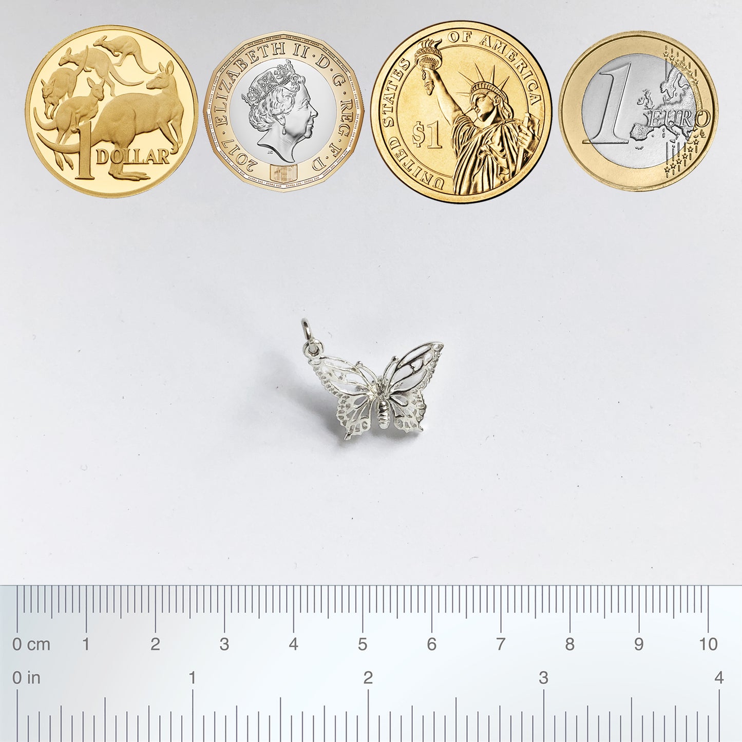 A silver filigree butterfly charm with coins and ruler for scale on a grey background.